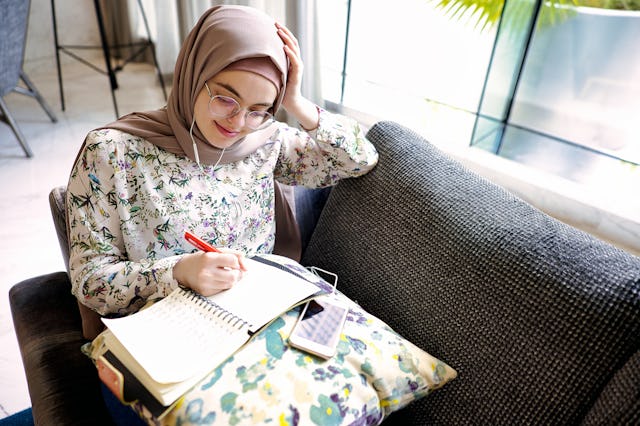 Woman sits in a booth near a window and listens to music while journaling for growth and reflection ...
