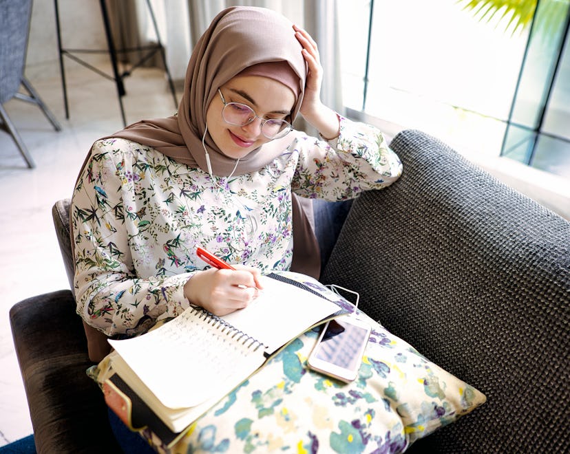 Woman sits in a booth near a window and listens to music while journaling for growth and reflection ...
