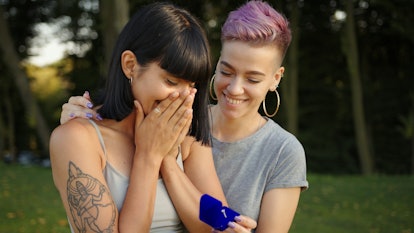 Woman holds engagement ring in front of another, who covers her mouth gasping with excitement