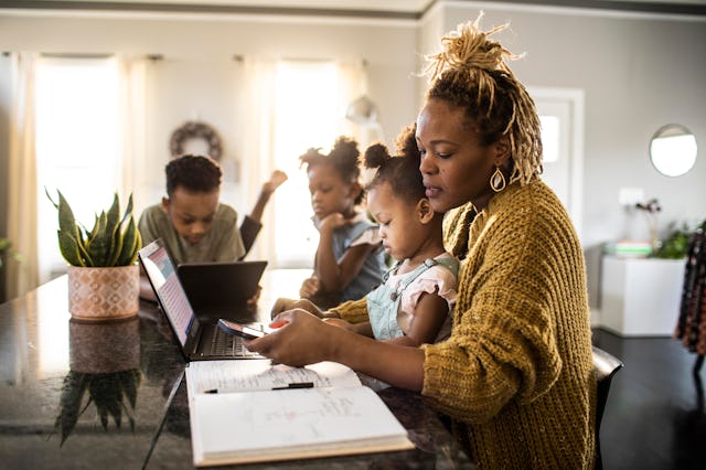 Mother working from home while holding toddler, family in background, like how a Libra prioritizes w...