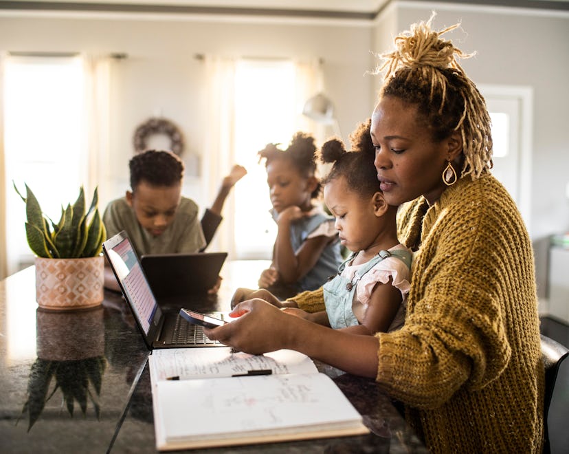 Mother working from home while holding toddler, family in background, like how a Libra prioritizes w...
