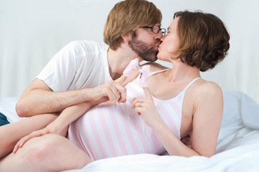 Cute expecting couple practicing playing with pigs puppets while kissing. White background.