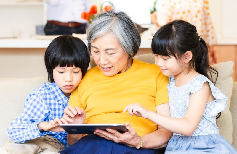 An elderly woman sharing a tablet computer with her two grandchildren as they look at something toge...