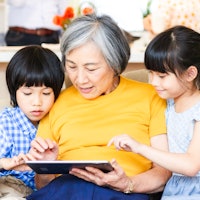 An elderly woman sharing a tablet computer with her two grandchildren as they look at something toge...