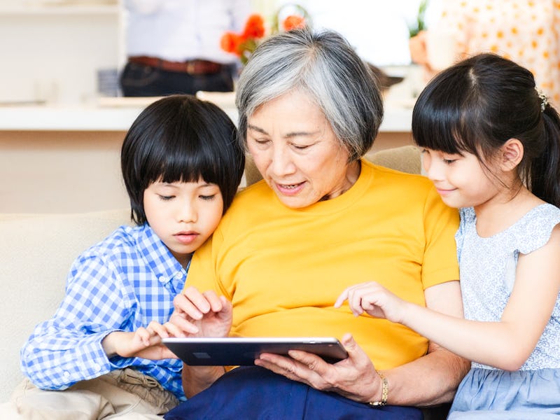 An elderly woman sharing a tablet computer with her two grandchildren as they look at something toge...