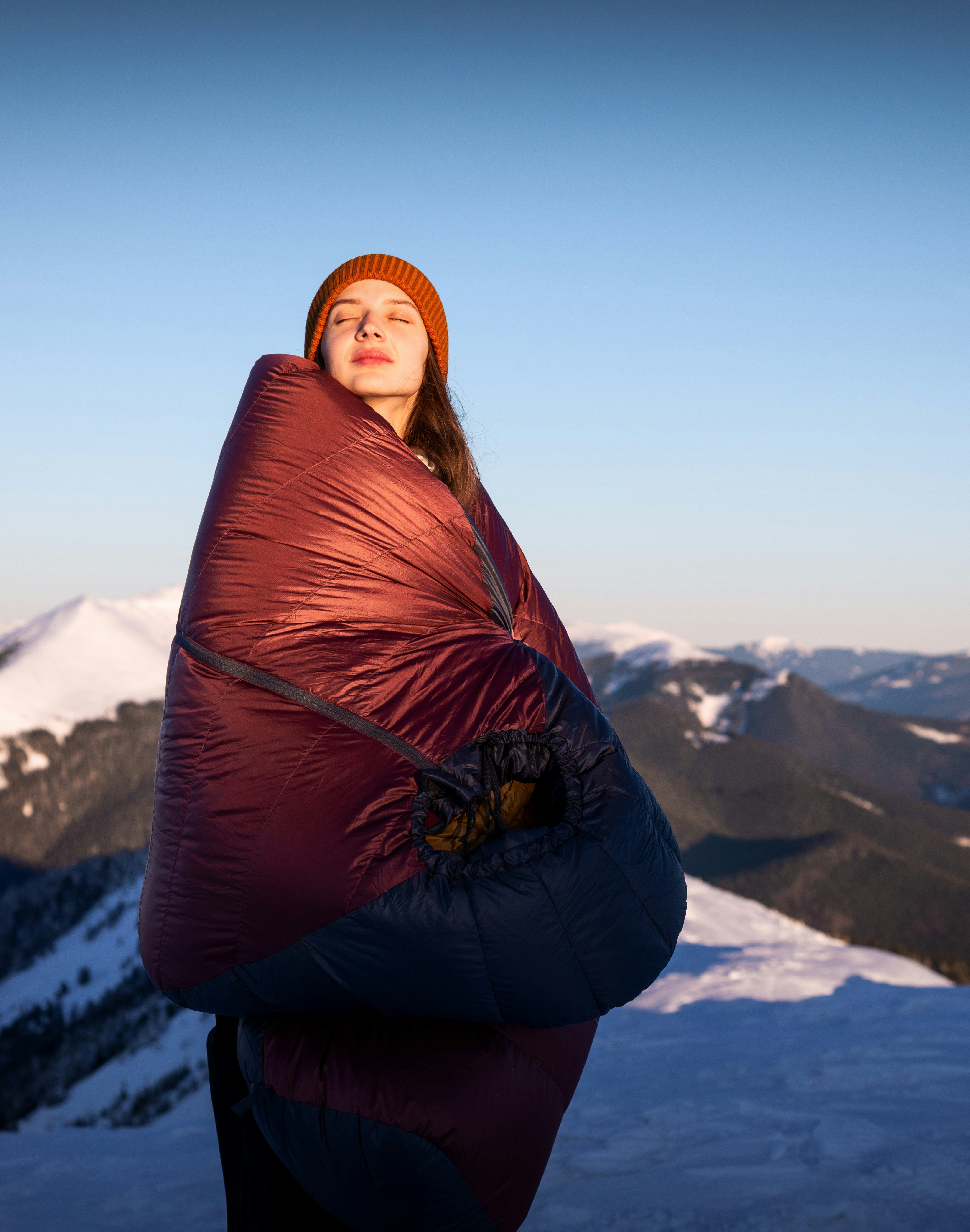 Smiling young woman staying in the background of a winter mountain wrapped in a warm sleeping bag. C...