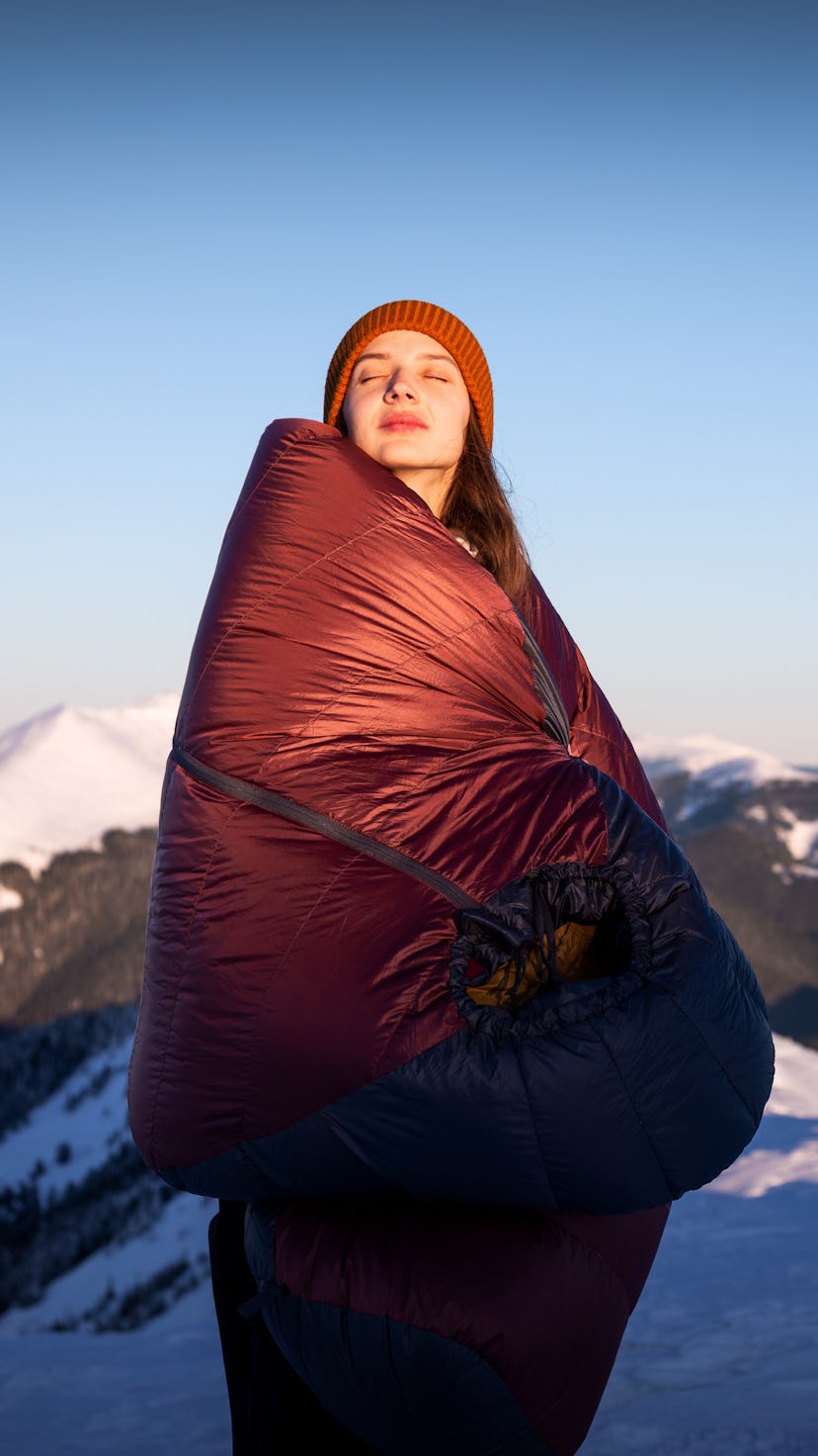 Smiling young woman staying in the background of a winter mountain wrapped in a warm sleeping bag. C...