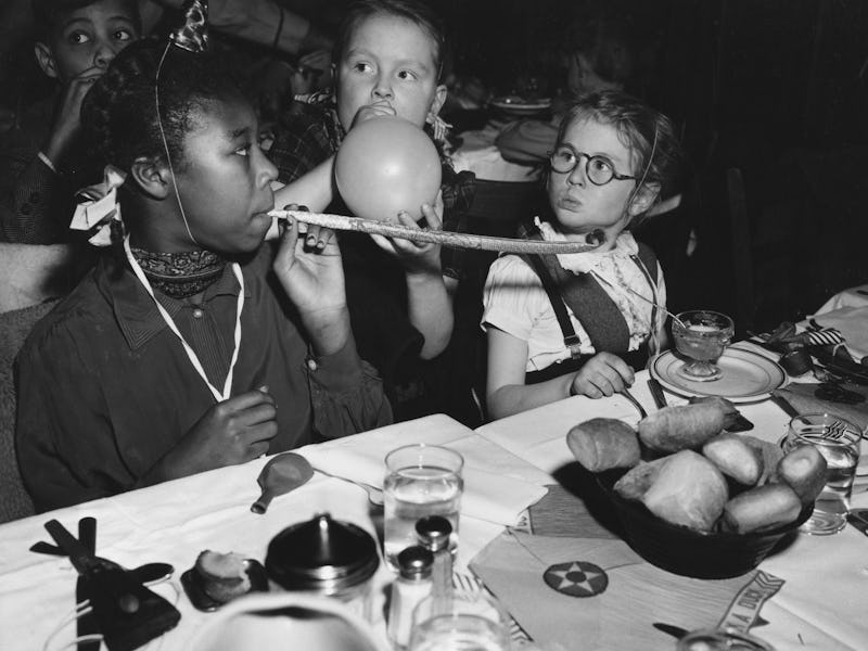 Lucille Carter blows a party whistle as Helen Green blows a balloon watched by Elizabeth Green, just...