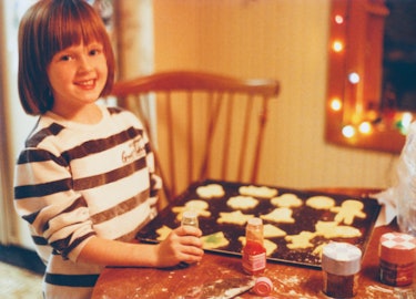 Vintage 1990s child baking Christmas cookies inside domestic kitchen. Happy child is decorating cook...