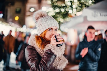 Woman in warm clothes and bobble hat having a mug of hot drink on Christmas market and looking at ca...