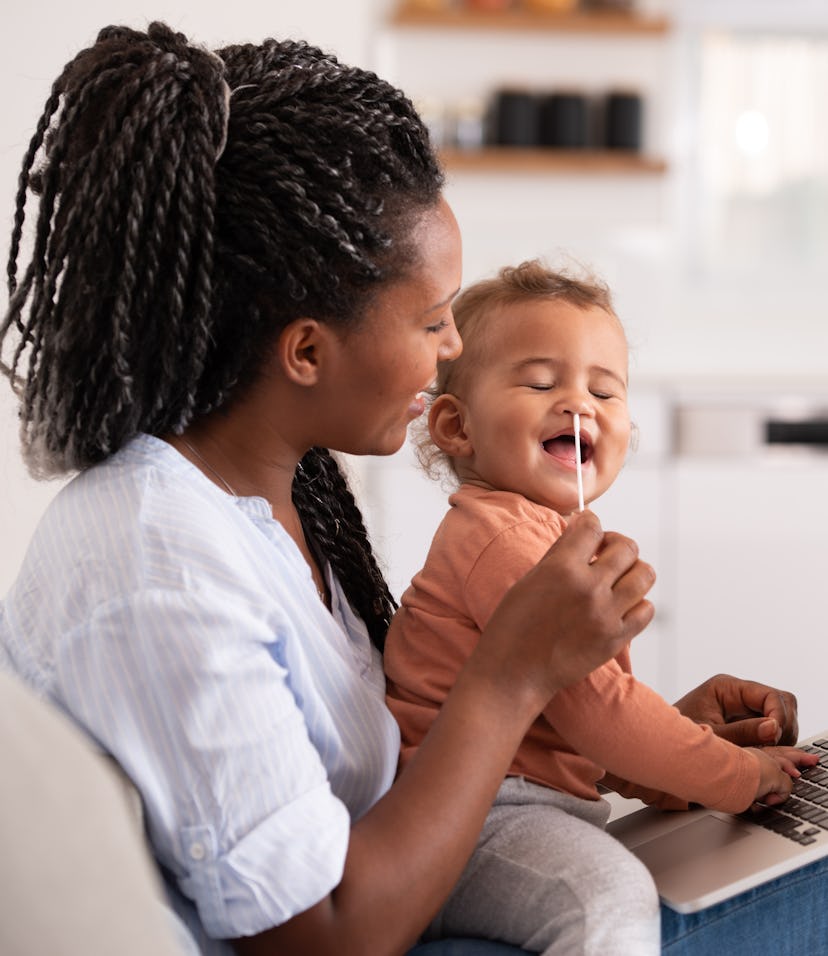 Woman using a swab of antigen self-test to test her baby for COVID-19, also using a laptop to commun...
