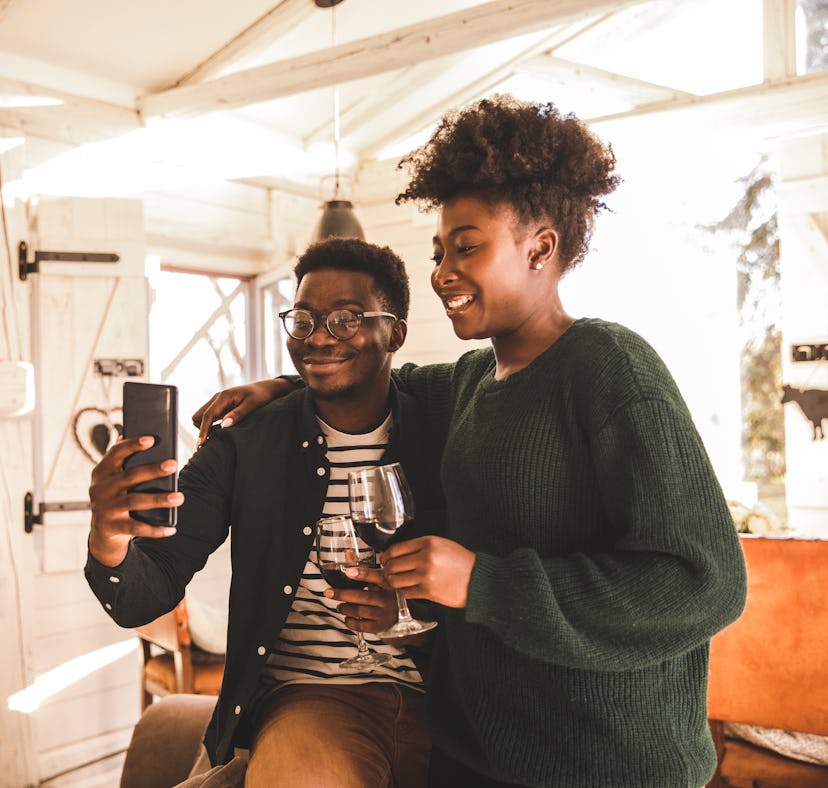 A young couple standing embraced in a cozy, rustic cabin, holding glasses of red wine and taking sel...