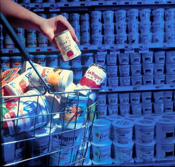 Hand depositing container of Yoplait A&P supermarket shelves into cart filled w. brands incl. Colomb...