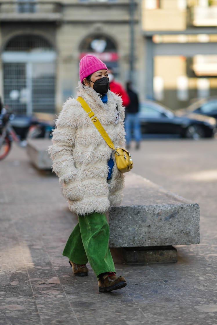 A guest colorblocks her pink beanie with a blue turtleneck, yellow bag, a white long coat, and green...