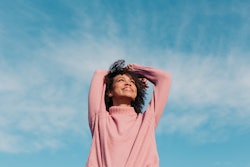 A woman wears a pink sweater against the sky. Here's your daily horoscope for February 15 2022.