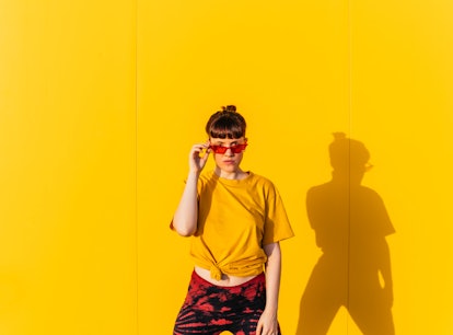young woman poses against yellow wall and holds her red glasses as she thinks about the emotional me...