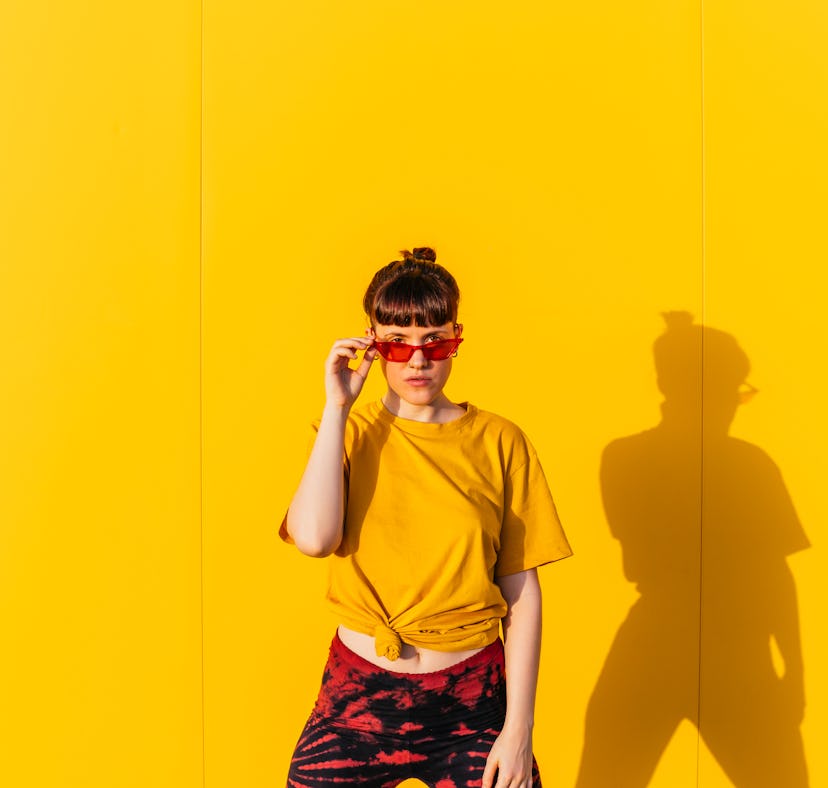 young woman poses against yellow wall and holds her red glasses as she thinks about the emotional me...