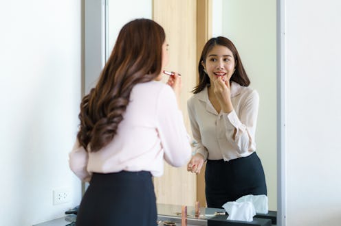 Beautiful Asian Woman wearing red dressed putting lipstick looking in mirror in her bedroom at home....