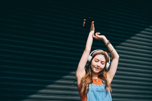 Carefree, smiling young Asian woman holding smartphone, dancing with her eyes closed while listening...