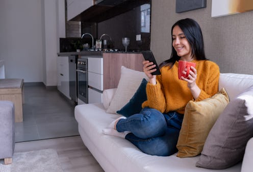 Beautiful young woman relaxing on couch texting on her smartphone while enjoying a coffee looking ve...