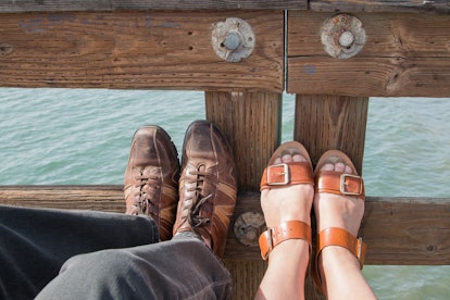 Two pairs of legs ( one man and one woman) with summer footwear resting their feet on a wooden raili...