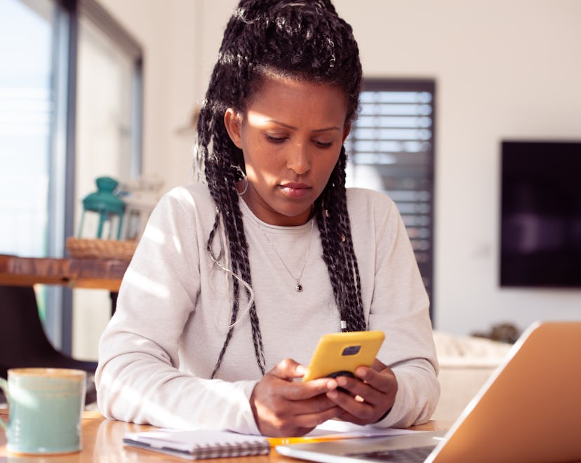 A businesswoman woman using a smartphone and laptop, sitting in the kitchen, drinking coffee. Female...