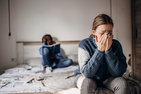 Stock image depicting woman crying on bed, her boyfriend is in the background.
