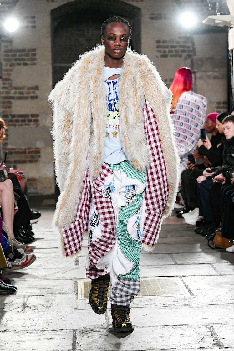 A model wearing a white shirt, green-white trousers and a beige fur and gingham coat by Matty Bovan