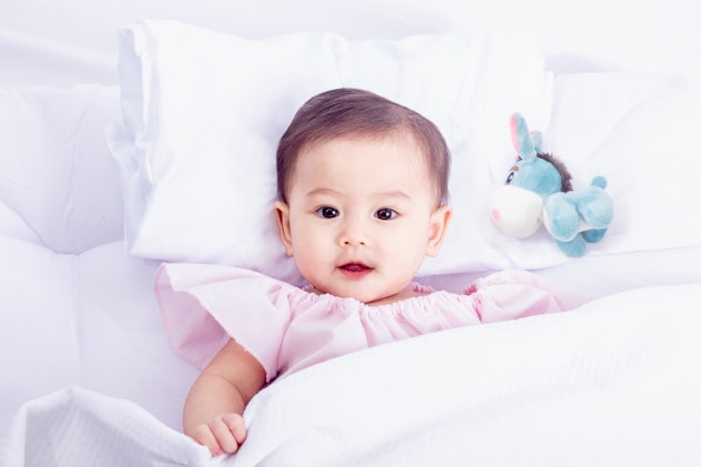 Toddler laying in bed on a white pillow wearing a pink dress, in a story about cute '90s baby names.