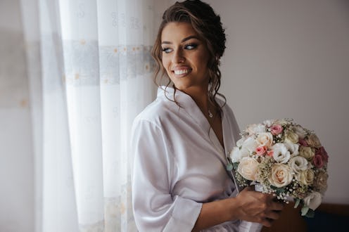 Beautiful young bride in white wedding dress holding bouquet and standing by the window at home, wai...