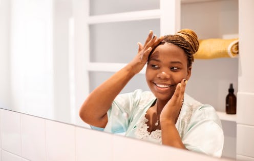 Beautiful young african woman touching face looking at skin in bathroom mirror