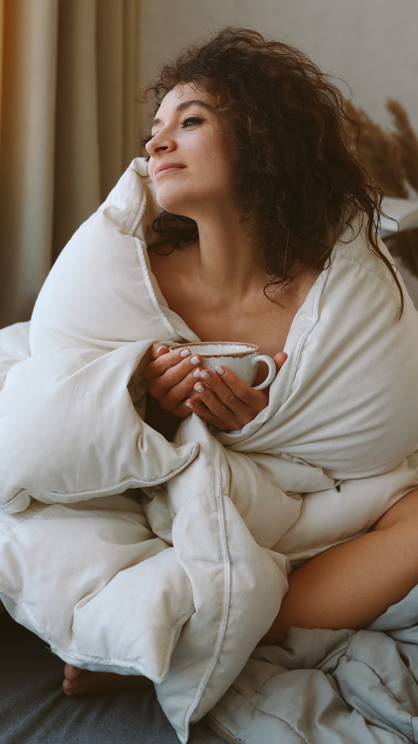 Relaxed young woman drinking coffee in bed and looking in window. The March 2022 new moon is station...