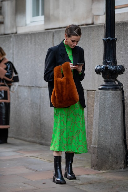 LONDON, ENGLAND - FEBRUARY 18: A guest is seen wearing brown bag, green dress, black blazer, ankle b...