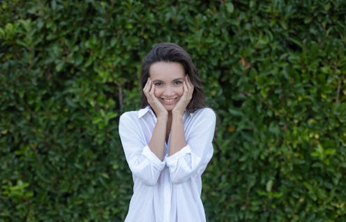 Portrait of smiling cute young woman with arms on cheeks in front of green plant wall.
