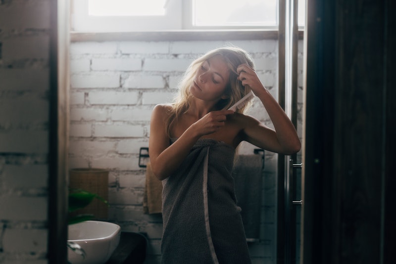 A young blonde woman standing in the bathroom wrapped in a gray towel, combing her hair.