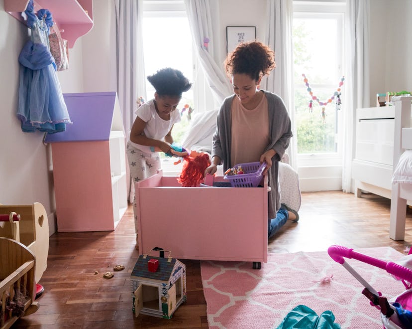 Mother and daughter picking up toys from container. Woman and girl are in bedroom. They are at home.