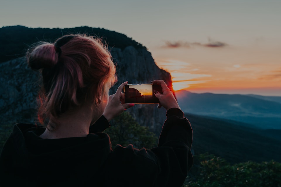 Young woman traveler hand holding smart phone and taking photo of majestic sunset in mountain during...