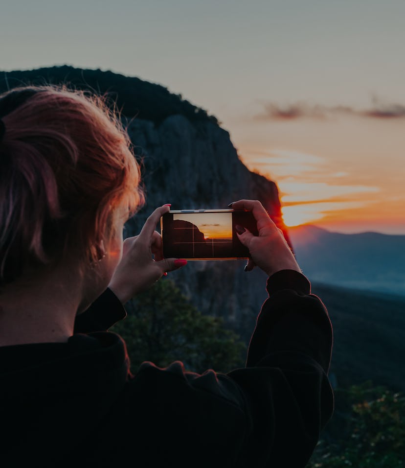 Young woman traveler hand holding smart phone and taking photo of majestic sunset in mountain during...