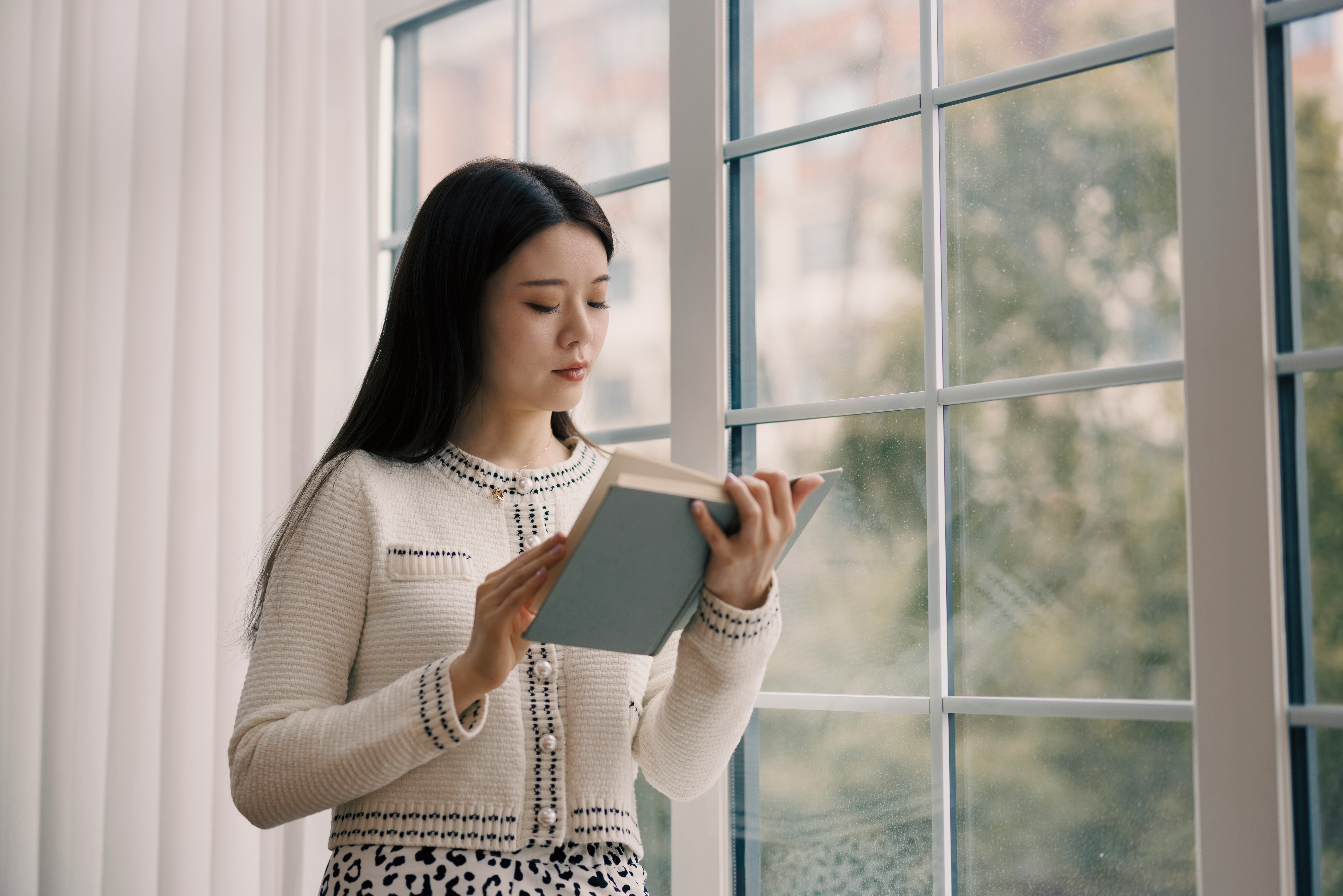 A young Asian woman, wearing a white sweater, stands by the window and reads a book.