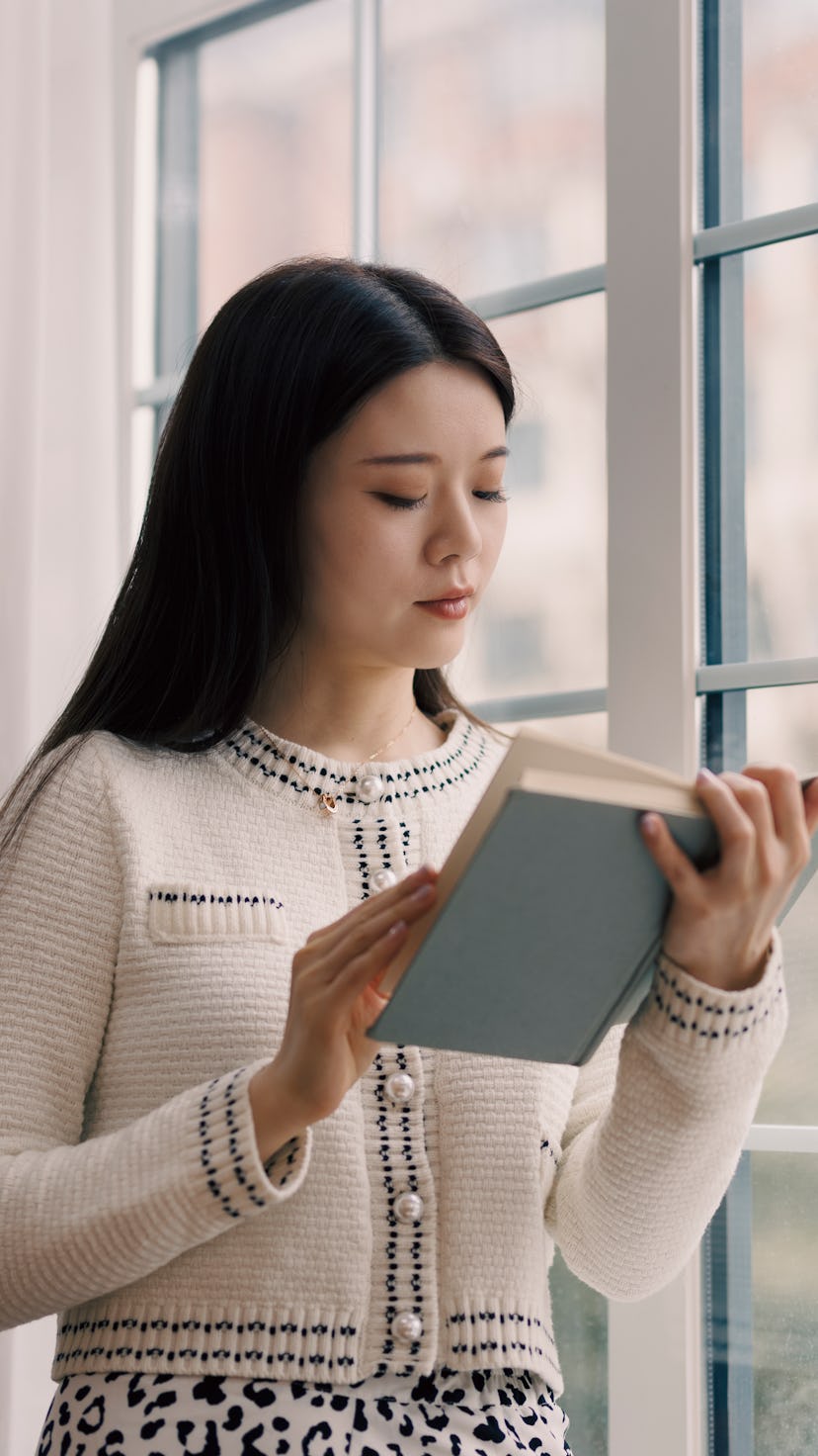 A young Asian woman, wearing a white sweater, stands by the window and reads a book.