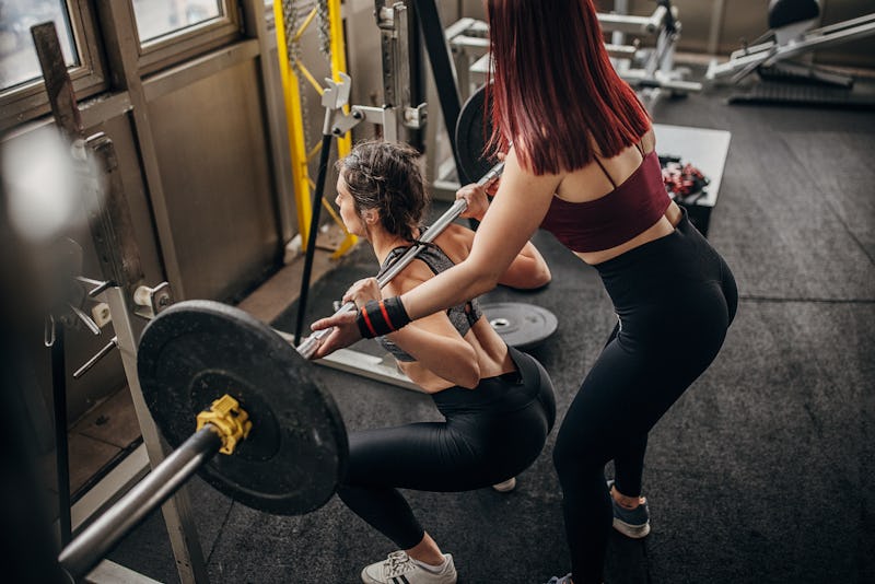 Two people, woman exercises with weights while her female personal trainer is assisting her in gym.