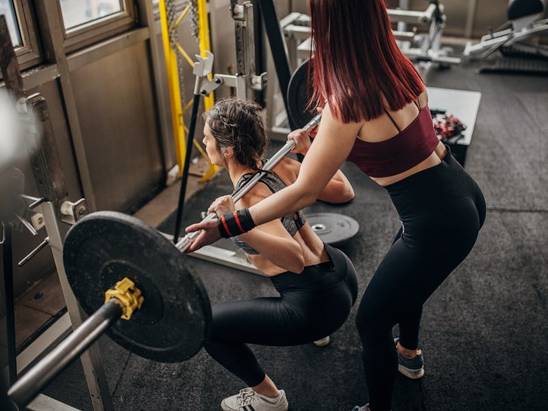 Two people, woman exercises with weights while her female personal trainer is assisting her in gym.