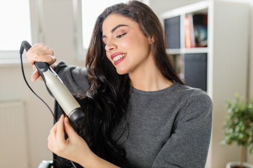 Woman looking at computer and using her curling iron while sitting at the dressing table. Woman does...