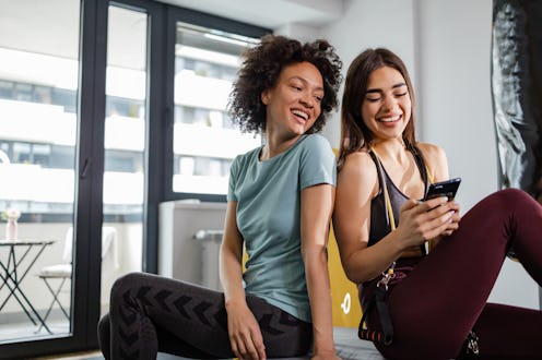 Photo of two woman taking a break from the exercise. They are looking at smart phone and laughing