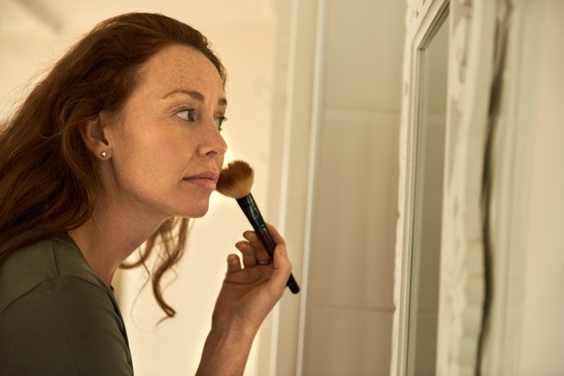 Young woman putting on a foundation with a make-up brush while standing in front of her bathroom mir...