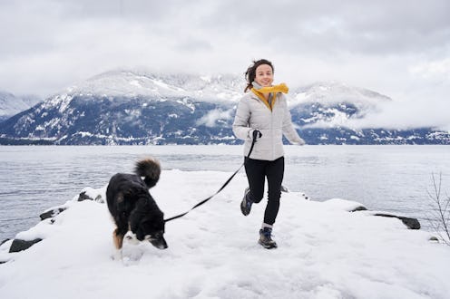 Smiling young woman running with her dog on a leash during a scenic walk by the ocean in winter