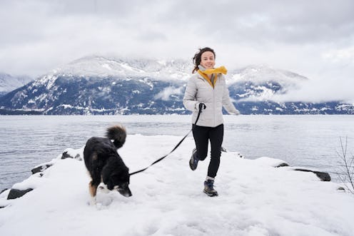 Smiling young woman running with her dog on a leash during a scenic walk by the ocean in winter