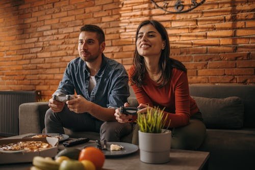 Couple sitting on sofa together and playing video games. These are the best video games for couples ...