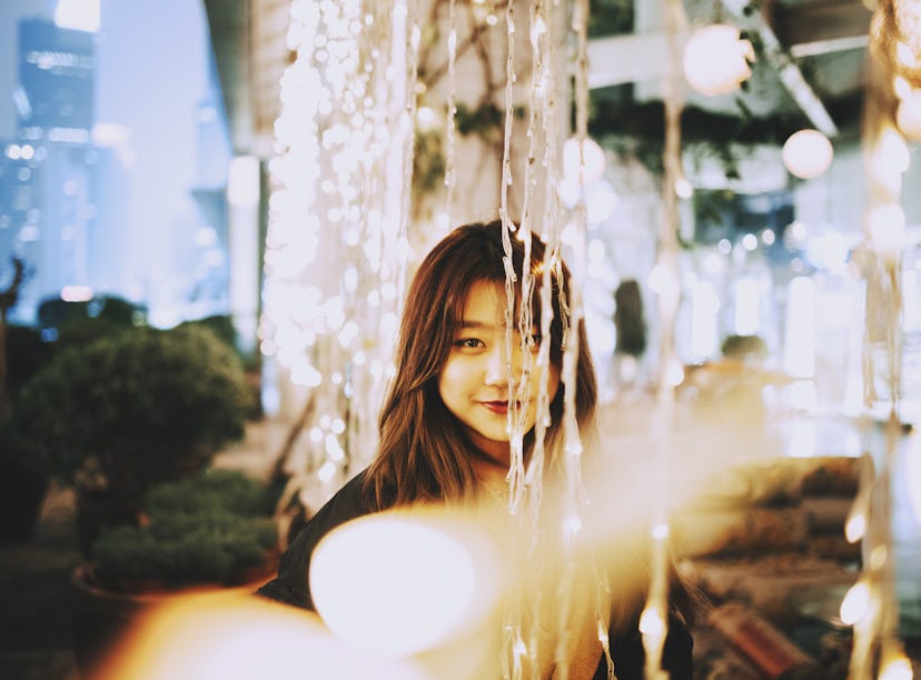 Young woman smiling through string lights, knowing her zodiac sign will be affected by the February ...