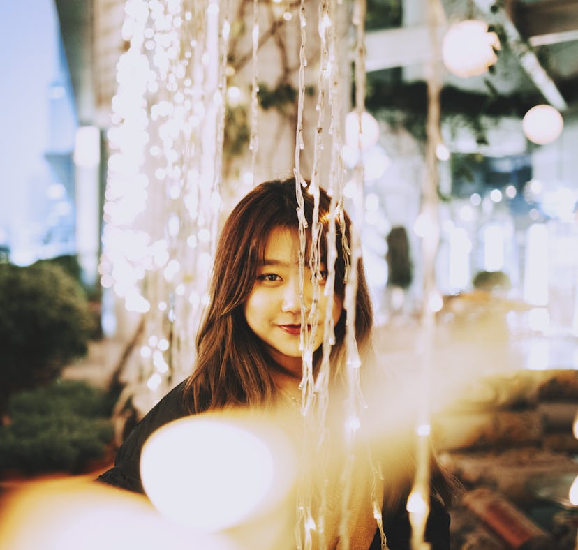 Young woman smiling through string lights, knowing her zodiac sign will be affected by the February ...